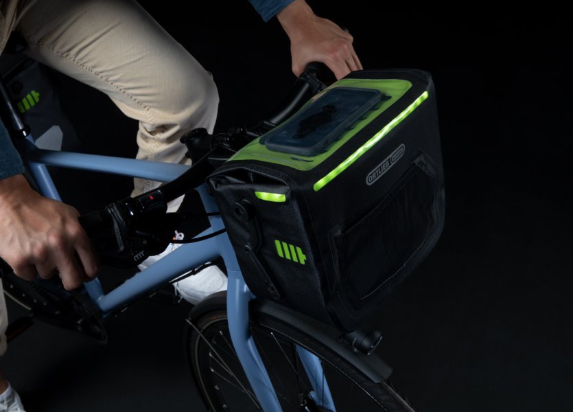 Ein Smartphone liegt auf einer schwarzen Fahrradtasche mit grünen Akzenten am Lenker. Die Tasche hat ein beleuchtetes, perforiertes Design. Der Hintergrund ist dunkel.