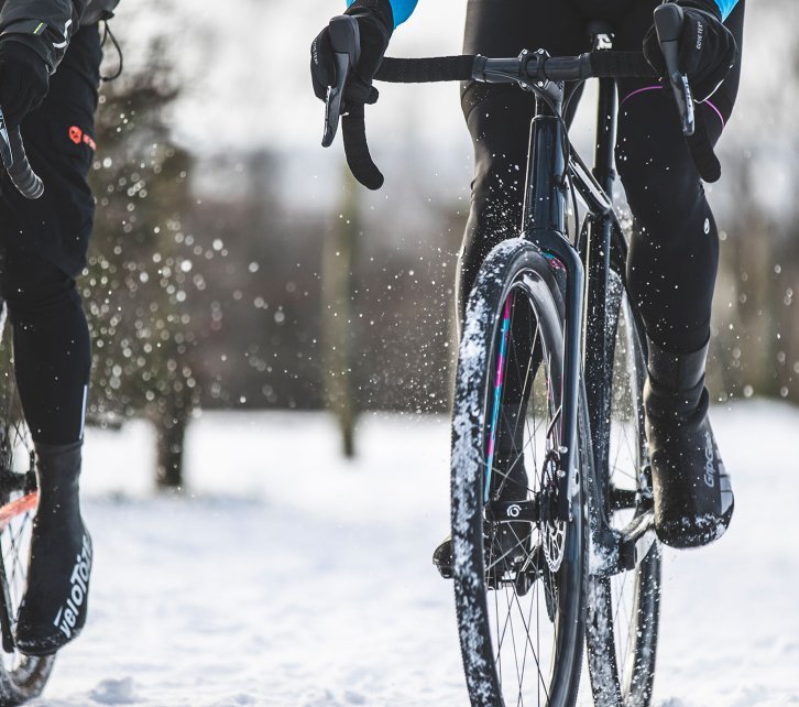 La photo montre deux cyclistes lors d'une sortie gravel dans la neige. L'image se concentre sur les vélos. Les deux cyclistes portent des surchaussures. 