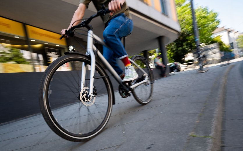 A man rides a bicycle dynamically through an urban environment, with a focus on the front wheel, while buildings and trees are blurred in the background.