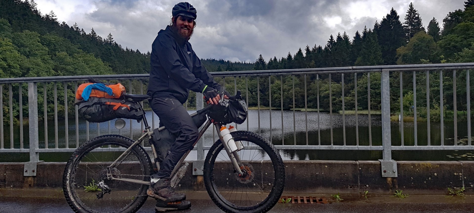 Ein Radfahrer mit Helm und Bikepacking-Taschen fährt auf einer nassen Brücke über einen Fluss, umgeben von dichtem Wald, bei bewölktem Wetter.