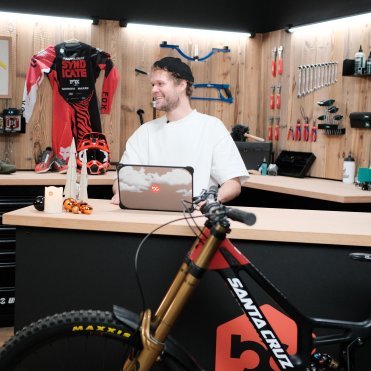 Escena en taller de MTB: persona en una mesa de trabajo con portátil, pared de herramientas al fondo y bicicleta Santa Cruz en primer plano.