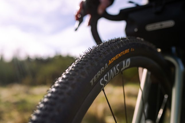 Close Up auf das Vorderrad eines Gravelbikes. Scharf im Vordergrund sehen wir Aufschrift und das grobe Profil an einem Continental Terra Adventure Reifenund den DT- Swiss Schriftzug