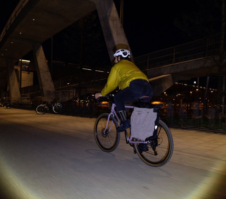Eine Frau auf einem Rennrad bei Nacht. Der weiße Helm und die retroreflektierende Radtasche sind gut erkennbar.