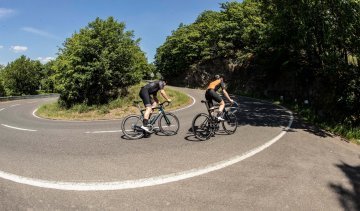 Two road cyclists, both on Specialized Tarmac SL7 bikes, are passing uphill on a winding road.