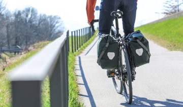 Ein Fahrradfahrer in einer roten Jacke fährt auf einem Radweg, mit zwei grünen Ortlieb Back-Roller Urban Fahrradtaschen, die an beiden Seiten des Hinterrads befestigt sind. Im Hintergrund sind eine grüne Wiese und kahle Bäume zu sehen.