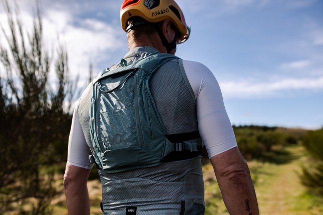 Ein Mann in Fahrradkleidung von schräg hinten, wir sehen nur den Oberkörper vor dem unscharfem Hintergrund einer Naturlandschaft mit grüner Vegetation und blauem Himmel. Der Mann trägt einen blaugrauen enganliegenden Trinkrucksack