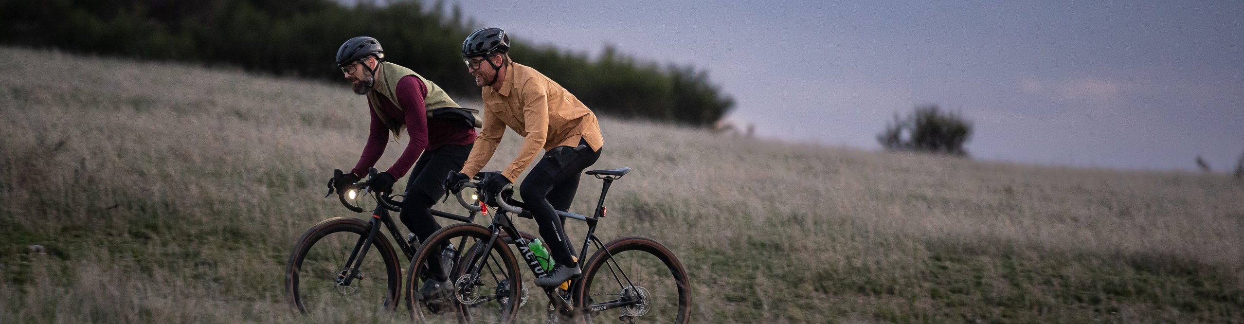 Zwei Radfahrer auf Gravelbikes fahren im Abendlicht durch eine Wiese, ausgestattet mit Helmen und Beleuchtung.