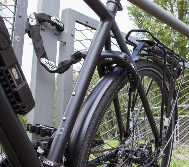 Bicicleta con candado de cuadro bloqueando la rueda trasera y una cadena adicional asegurada a una valla metálica.