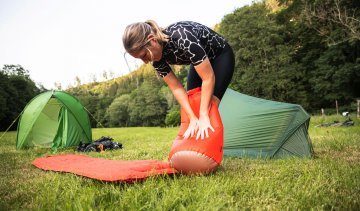 2 VAUDE tents pitched on a meadow surrounded by trees. In the foreground, a pump bag is used to fill a camping mat with air.