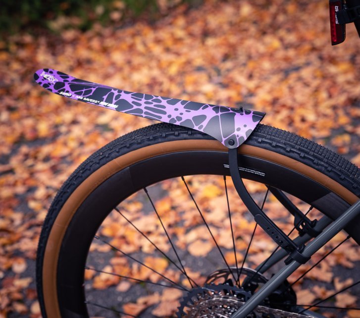 A black Ass Savers "Win Wing" mudguard with a purple splatter design mounted on the rear wheel of a grey bicycle.