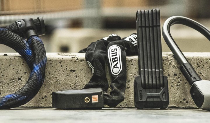 Mehrere Fahrradschlösser – Kabelschloss, Kettenschloss, Faltschloss und Bügelschloss – liegen nebeneinander auf einem Betonblock in urbaner Umgebung.