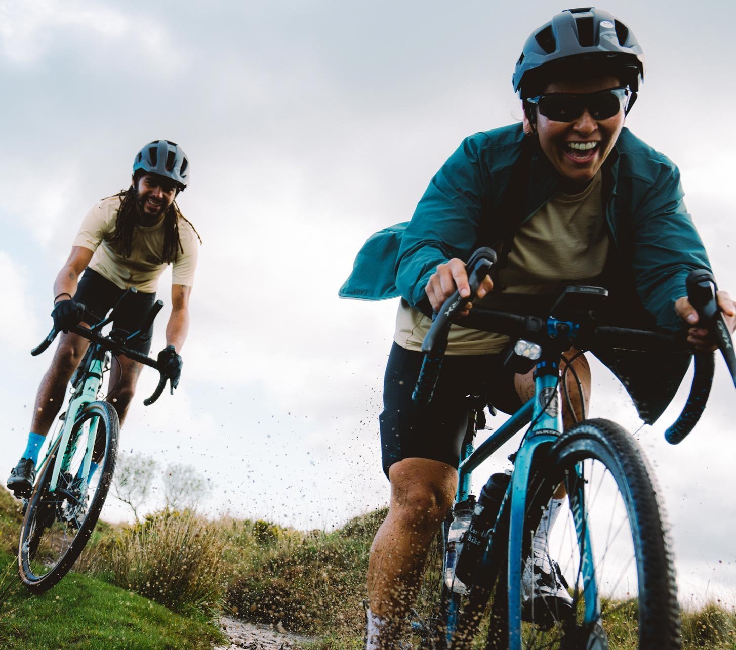 Deux cyclistes gravel avec casque roulent rapidement sur un chemin de gravier mouillé en éclaboussant de l’eau, à vélo gravel avec des vêtements Endura dans un paysage ouvert.