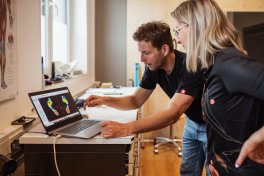 Zwei Personen stehen an einem Tisch und betrachten ein 3D-Modell eines Fahrradsattels auf einem Laptop-Bildschirm, während sie die Anpassung diskutieren.