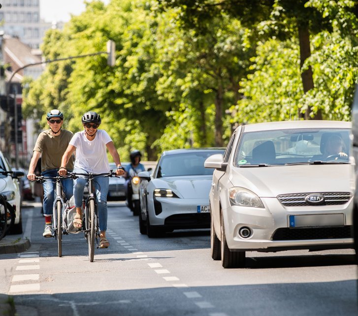 Two people are riding bicycles on a bike lane in a city street, while cars are stuck in traffic next to them. Trees and buildings are visible in the background.