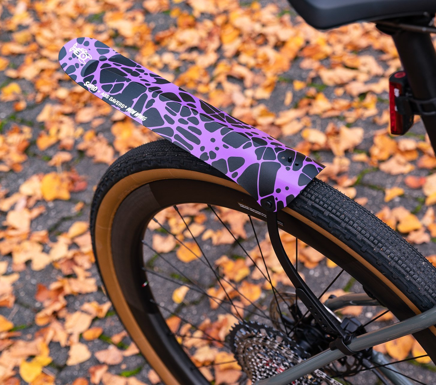 Ein leichtes "Win Wing"-Schutzblech der Marke Ass Savers in schwarz mit Lila Splatter-Muster am Hinterrad eines grauen Gravel-Bikes. Im Hintergrund Herbstlaub