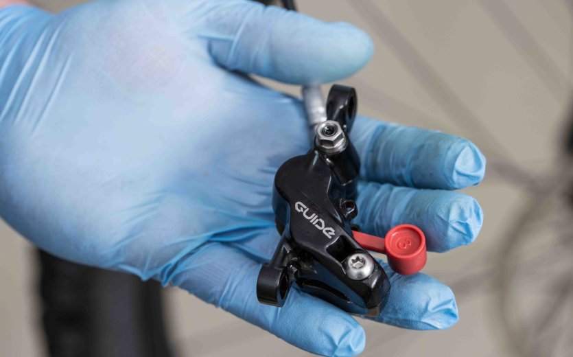 Eine Hand mit blauen Handschuhen hält eine Bremszange der hydraulischen Scheibenbremse mit einer roten Entlüftungsschraube, bereit zur Wartung, mit einem Fahrrad im Hintergrund.