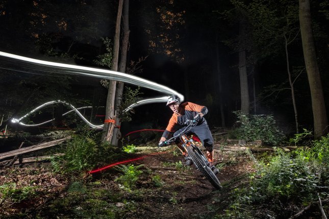 Un ciclista de montaña pedalea de noche en un sendero del bosque, iluminado por una luz en el casco, con trazos de luz marcando el camino y árboles rodeando el entorno.