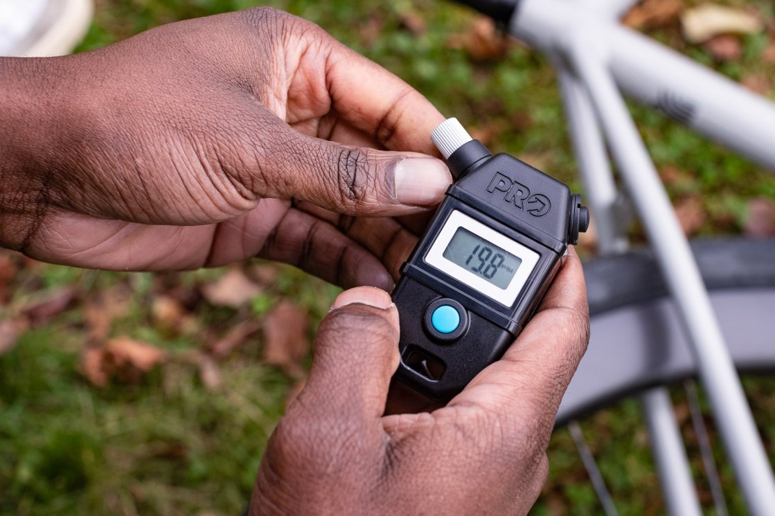 Zwei Männerhände halten einen Pro Luftdruckprüfer. Im Hintergrund liegt ein Fahrrad auf einer Wiese