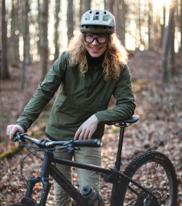 bc employee Kai leans casually against his MTB in the Aachen forest, smiling. Kai has long blonde curls and wears a light-colored MTB helmet and an olive rain jacket.