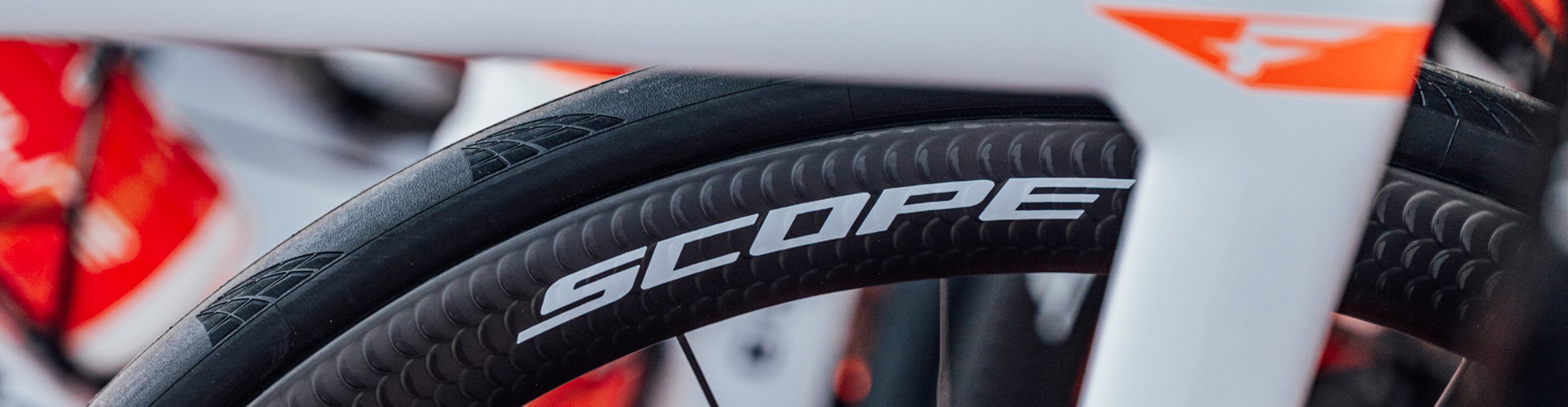 Close-up of a Scope Cycling carbon wheel on an INEOS Grenadiers road bike with a white and red frame and black aero rim.
