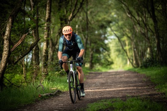 Cycliste sur un vélo gravel en forêt, mains posées sur les leviers de frein et de vitesse (STI), adoptant une position légèrement redressée, équipé d’un casque et d’une tenue de cyclisme.