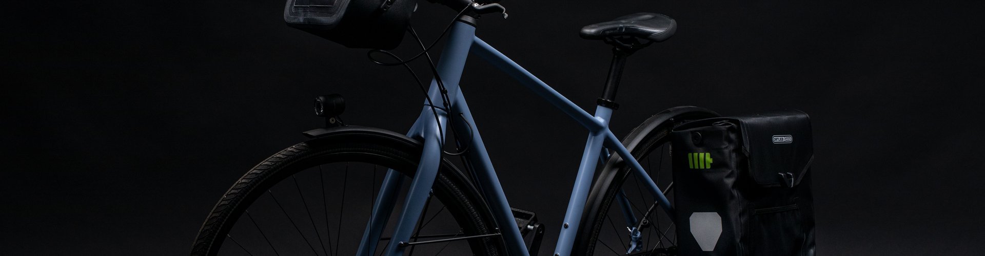 Ein blaues Fahrrad mit einer schwarzen ORTLIEB Lenkertasche und einer Gepäcktasche hinten, beide mit grünen Akzenten. Die Lenkertasche hat ein beleuchtetes Design. Der Hintergrund ist dunkel.