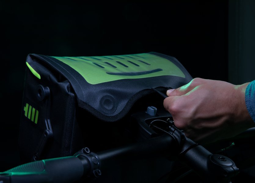 Eine Hand öffnet eine schwarze Fahrradtasche mit grünen Akzenten. Die Tasche hat ein beleuchtetes, perforiertes Design. Der Hintergrund ist dunkel.