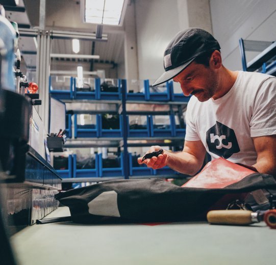 Andi à l'un des postes de travail de l'atelier chez ORTLIEB. Il tient un fermoir de sacoche dans la main. Devant lui se trouve une sacoche à bagages.