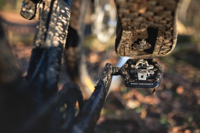 Detail photo: The sole of an MTB shoe, positioned just above a black OneUp Clip Pedal. In the blur, parts of a dark mountain bike and an autumn forest floor are visible.