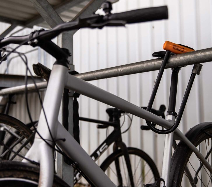 Bicicleta asegurada con un candado en U al cuadro y a una barra metálica en un aparcamiento cubierto.