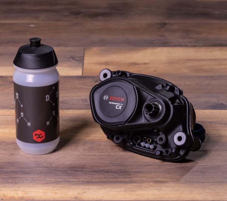 A Bosch Performance CX e-bike drive lies on a dark wooden floor. Next to it is a bike water bottle