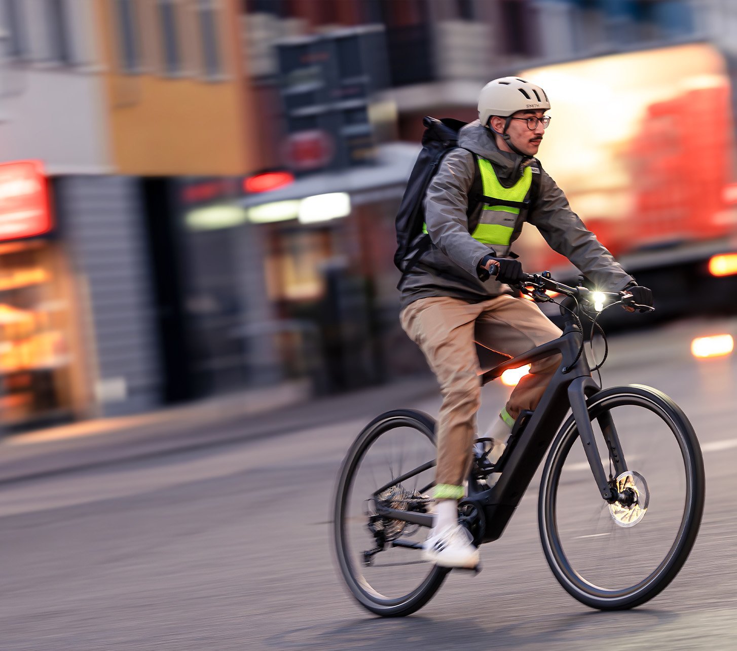 Pendler fährt mit E-Bike durch die Stadt, mit Helm, Rucksack und Beleuchtung im Straßenverkehr, nachhaltige Alternative bei hohen Benzinpreisen