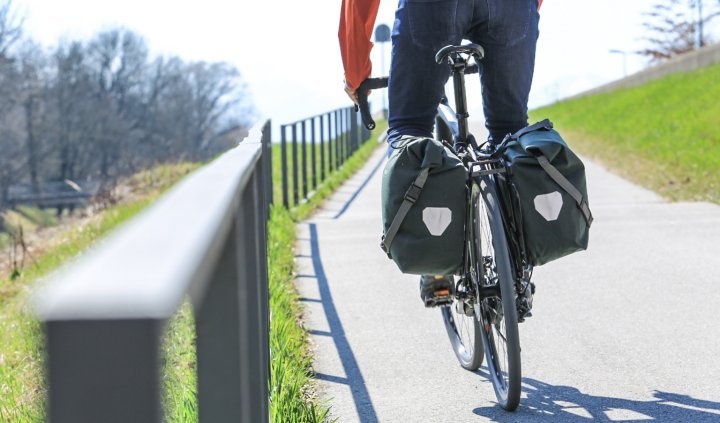 Un cycliste en veste rouge roule sur une piste cyclable, avec deux sacs de vélo Ortlieb Back-Roller Urban verts fixés de chaque côté de la roue arrière. Une prairie verte et des arbres dénudés sont visibles en arrière-plan.