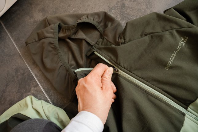 Eine grüne Funktionsjacke liegt auf einem grau gefliesten Fußboden. Eine Hand zieht den Frontreißverschluss zu.
