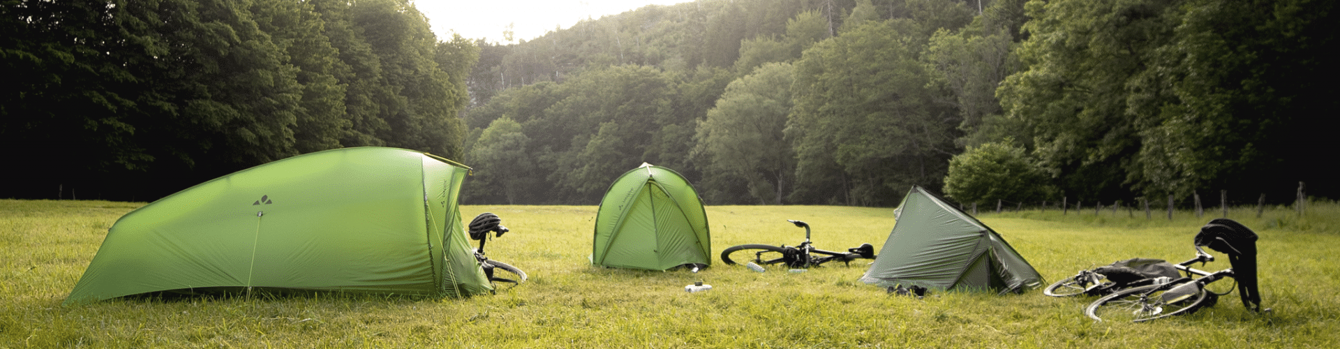 3 VAUDE tents pitched on a meadow surrounded by trees. Next to each tent is a bicycle.