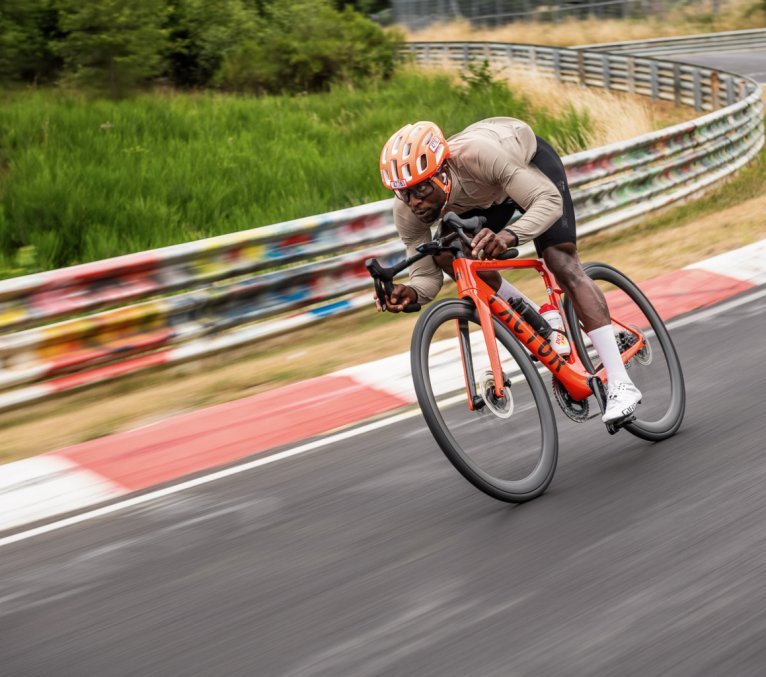 Ein sportlicher, junger Mann mit orangem Helm fetzt auf einem roten Rennrad über eine Auto-Rennstrecke