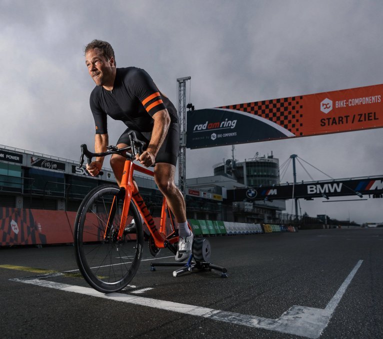 A man on an orange road bike trains on a smart trainer at the start line of the Nürburgring racetrack during the “Rad am Ring” event.