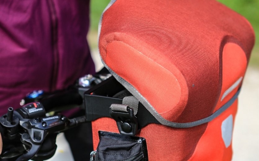 A red Ortlieb bicycle bag is attached to a bike. In the foreground, part of the handlebar with shifters and a hand are visible, while a blurry green field can be seen in the background.