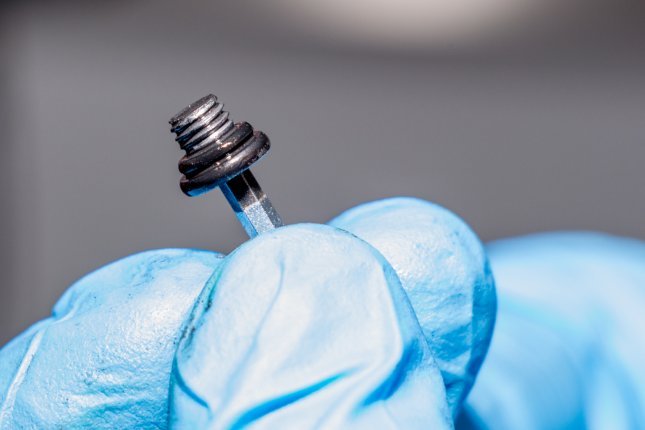 A hand wearing blue gloves holds a bleed screw with a black seal to check it, with a blurred background.