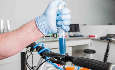 Eine Einwegspritze, halbvoll mit einer klar-blauen Flüssigkeit am Bremshebel eines Fahrrads beim Entlüften des hydraulischen Systems