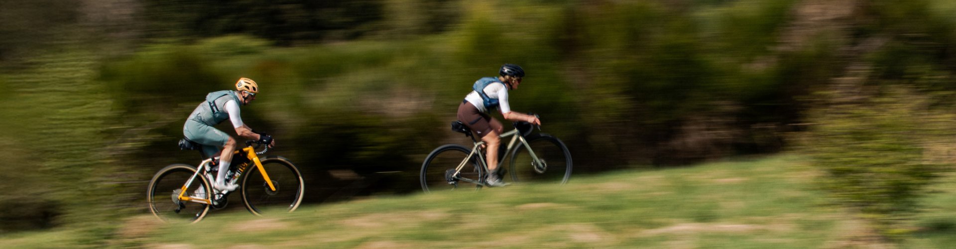 Herbert und Katrin flitzen auf ihren Gravel Bikes durch eine grüne Naturlandschaft. Mitzieheraufnahme von der Seite, nur die beiden Radfahrenden sind scharf abgebildet, der Hintergrund verwischt.