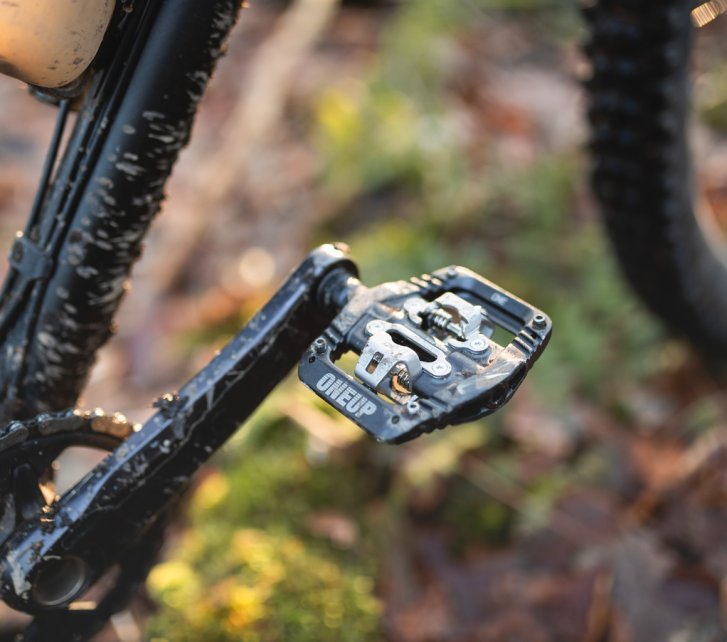 Detail photo: A black OneUp Clip Pedal on a dark mountain bike. We only see a crank arm and parts of the down tube. In the blurred background, an autumn forest floor and a partially visible front wheel can be seen.