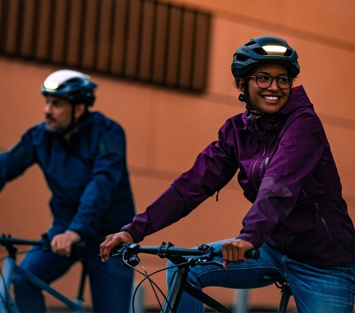 Dos ciclistas con cascos que tienen luces integradas circulan por la ciudad al anochecer. Llevan ropa adecuada para el clima y son muy visibles en el tráfico.