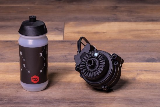 A TQ HPR50 e-bike drive lies on a dark wooden floor. A bike water bottle stands next to it
