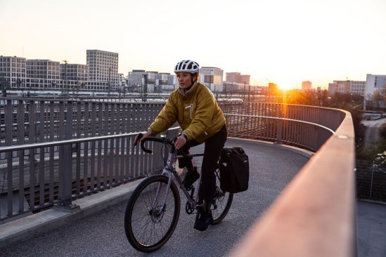 Eine Frau auf einem Rennrad fährt über eine Bahnüberführung. Dahinter sehen wir Bürohäuser und Bahngleise im Gegenlicht der untergehenden Sonne. Die Frau trägt einen weißen Helm, eine ockerfarbene warme Jacke hinten am Rad ist eine schwarze Panniertasche.