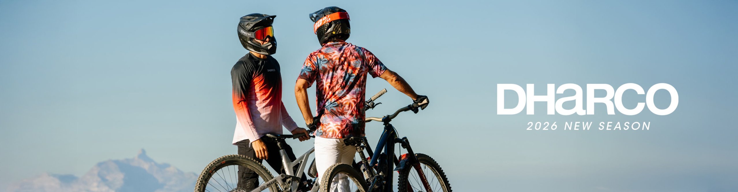 Zwei Mountainbike Fahrer stehen mit ihren MTB's vor einem wolkenlosen Alpenpanorama Hintergrund und tragen auffällige und stylische Fahrradbekleidung von DHaRCO