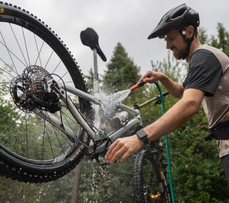 Mann mit Helm reinigt ein Mountainbike im Freien mit einem Wasserschlauch. Das Hinterrad ist angehoben, Wasser spritzt auf Rahmen, Antrieb und Kassette. Das Fahrrad wird gründlich abgespült, um Schmutz und Rückstände zu entfernen.