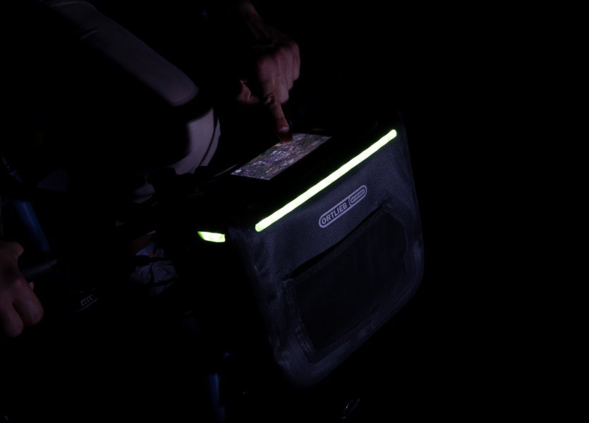 Eine Hand berührt ein Smartphone auf einer schwarzen Fahrradtasche mit grünen Akzenten am Lenker. Die Tasche hat ein beleuchtetes, perforiertes Design. Der Hintergrund ist dunkel.