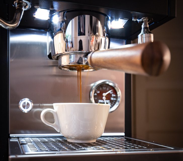 Eine klassische, cremeweiße Cappucchinotasse steht unter einem bodenlosen Siebträger, aus dem nussbraune Crema tropft. 