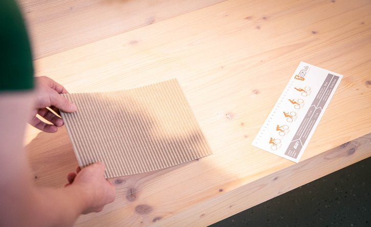 Two hands place a rectangular piece of corrugated cardboard on a light wooden bench. Next to it lies a measuring gauge from SQlab.